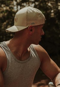 Világos bézs színű baseball sapka perforált mintával, kis logóval és ívelt szélekkel. Szürke ujjatlan pólóval viselve.