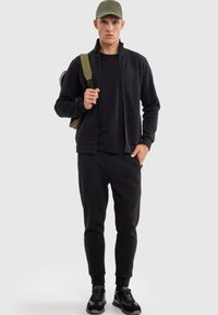 Giacca nera con zip, t-shirt nera e pantaloni jogger affusolati. L'outfit ha una texture liscia, con un cappellino verde e uno zaino grigio. Le sneakers nere completano il look.
