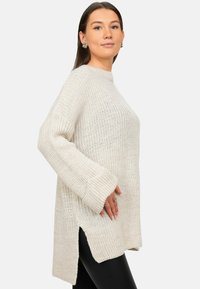 Overmaat beige gebreide trui met een geribbelde nek, zijsplitten en brede mouwen, gemaakt van textuurgaren voor een relaxte pasvorm.