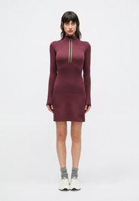 Långärmad klänning i vinrött med hög krage, utrustad med ett centralt blixtlås med en mångfärgad rand. Mjuk, elastisk tyg. Matchas med vita sneakers.