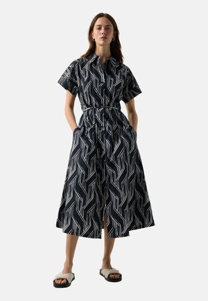 Kvinde stående iført sort og hvid midi-kjole med geometrisk mønster, korte ærmer, knaplukning foran, bånd i taljen og hvide slip-on sandaler.