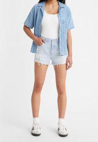 Chemise en denim bleu clair à manches courtes, débardeur blanc, shorts en denim bleu clair déchirés, associés à des baskets blanches avec des accents noirs.