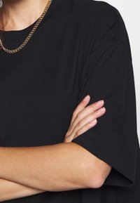 T-shirt en coton noir avec une coupe ample, des manches courtes et un col rond. Présente un collier en chaîne dorée porté avec les bras croisés.