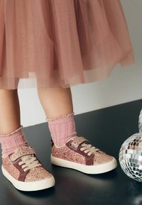 Glitzernde pinke Sneaker mit einem braunen Wildlederakzent, beigen Schnürsenkeln und einem Klettverschluss; getragen mit gerippten pinken Knöchelsocken, auf einer dunklen Oberfläche.