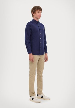 Camicia a maniche lunghe blu navy con colletto, dotata di bottoni e piccolo logo, abbinata a pantaloni beige e sneakers bianche con dettagli neri.