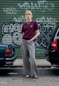 Burgunderfarget grafisk T-skjorte, løse grå denimbukser med vide ben og lyse sko. Bakgrunnen har grønn metall og urbane graffiti.