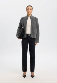 Cárdigan gris de mezcla de lana con cuello alto y bolsillo delantero, combinado con una blusa crema, jeans negros y tacones negros puntiagudos, sosteniendo un bolso negro.