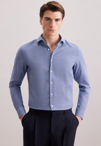 Man in een lichtblauw gestreept overhemd met lange mouwen en donkere broek, met zijn handen in zijn zakken, staand tegen een effen achtergrond.