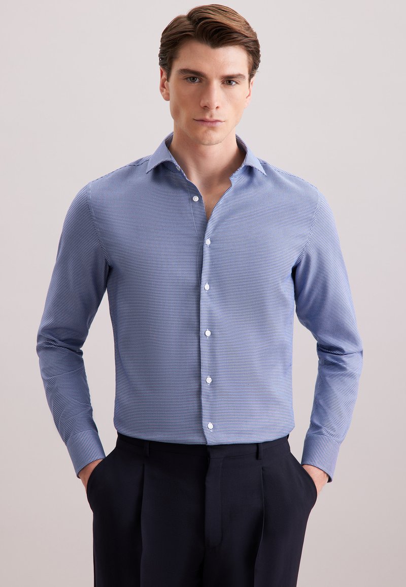 Man in een lichtblauw gestreept overhemd met lange mouwen en donkere broek, met zijn handen in zijn zakken, staand tegen een effen achtergrond.