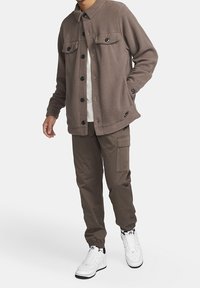 Brun fleecejacka med två bröstfickor, svarta knappar och krage, bärs med olivgröna byxor och vita sneakers.