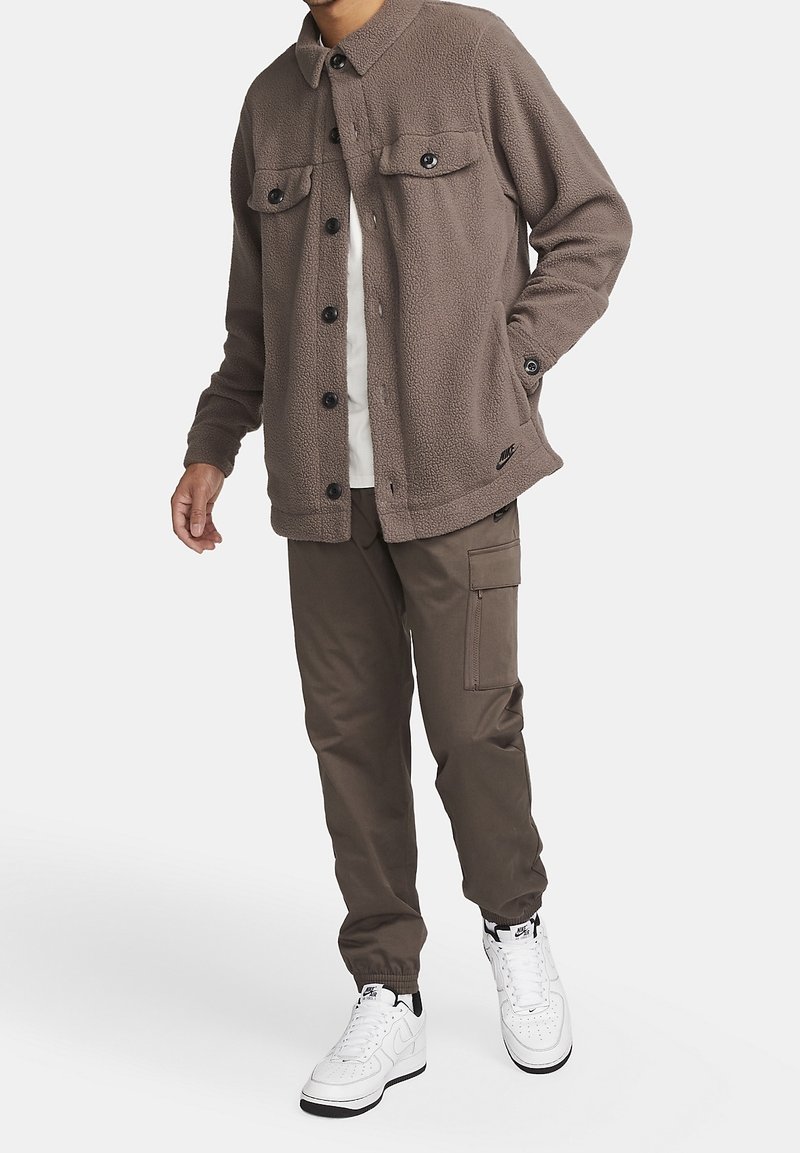 Brun fleecejacka med två bröstfickor, svarta knappar och krage, bärs med olivgröna byxor och vita sneakers.