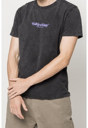 Zwarte katoenen t-shirt met een "Take it Easy" graphic in paars. Korte mouwen, ronde hals. G draagt het met lichtbeige shorts.