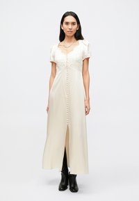 Lange, crèmekleurige jurk met kanten lijfje en pofmouwen. Heeft een knoopsluiting aan de voorkant en een splitsing vooraan. Gecombineerd met zwarte laarzen.