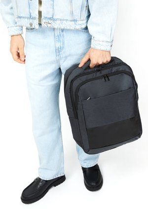 Black and gray backpack made of fabric with a smooth texture, featuring a zippered front pocket and top handle. Worn by a person in denim jeans.