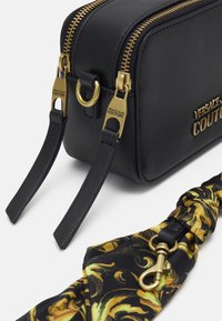 Borsa in pelle nera con zip e hardware dorato, caratterizzata da un logo prominente, affiancata da una tracolla in tessuto stampato in nero e oro.