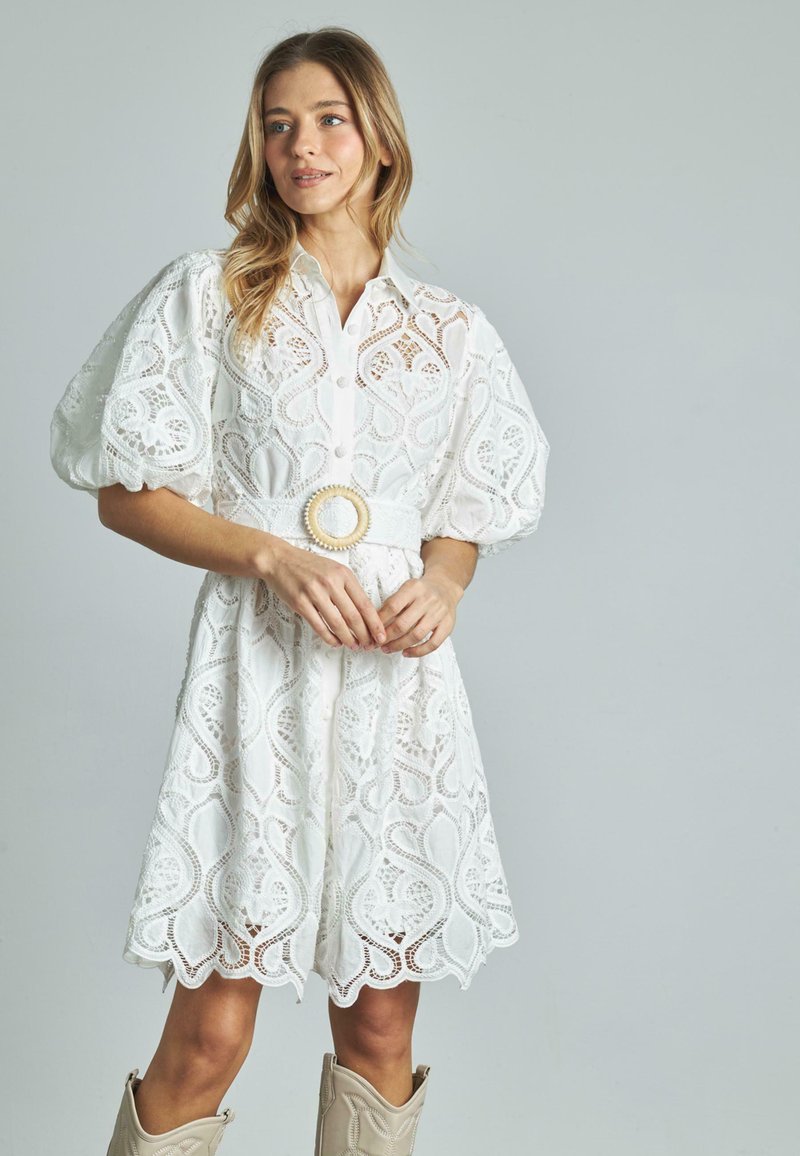 Biała koronkowa sukienka z zapięciem na guziki z przodu, bufkowymi rękawami i dekoracyjnym paskiem, z falbaniastym dołem oraz skomplikowanymi wzorami wyciętymi.