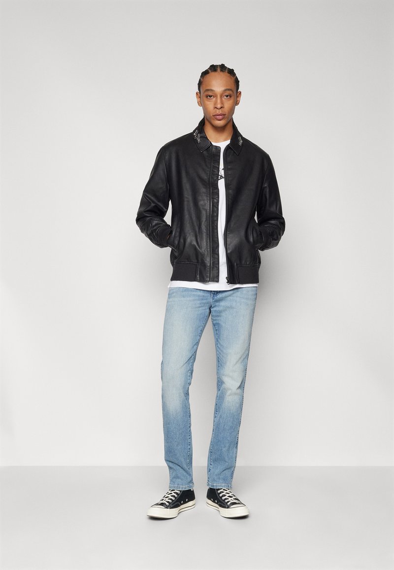 Mężczyzna z warkoczami, ubrany w czarną skórzaną kurtkę, białą koszulkę, jasnoniebieskie dżinsy i czarne trampki, stojący przy gładkiej ścianie.