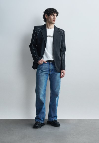 Νέος άνδρας όρθιος, φοράει μαύρο σακάκι με λευκές λεπτομέρειες, λευκό μπλουζάκι με γραφικό σχέδιο, μπλε τζιν και μαύρα αθλητικά παπούτσια, κοιτάζοντας προς το πλάι.