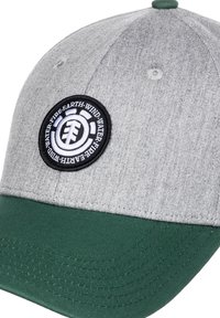 Czapka baseballowa z jasnoszarym wierzchem i ciemnozielonym daszkiem, z czarną i białą okrągłą naszywką z napisem: "Ziemia, Wiatr, Woda, Ogień" wokół logo drzewa.