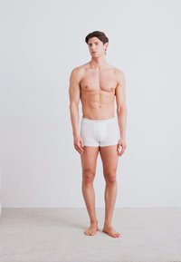 Weiße Baumwollboxershorts an einem männlichen Model, das auf einem hellen Teppich steht. Die Shorts haben ein einfaches Design mit einem weichen Bund.