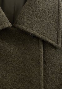 Primo piano di un cappotto di lana marrone con un ampio colletto e un bottone nero visibile vicino allo scollo.