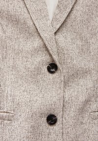 Hellgrauer strukturierter Blazer mit einem dezenten Muster, ausgestattet mit zwei schwarzen Knöpfen und Reverskragen. Die Konstruktion umfasst Schubtaschen.