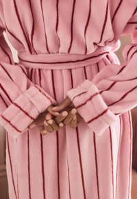 Pink fleece robe with red vertical stripes, featuring wide sleeves and a tie belt. Hands clasped together in front, showcasing natural nails.