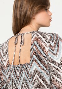 Blusa impressa com padrão em chevron nas cores castanho, azul e branco. A parte de trás tem um recorte quadrado com um fecho de laço. Tecido leve, mangas bufantes.