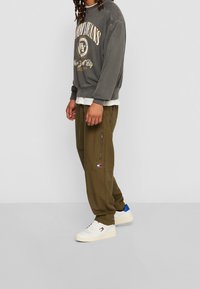 Felpa grigia con logo, indossata sopra una maglietta bianca a strati, abbinata a pantaloni cargo oliva con zip e sneakers bianche con dettagli blu.