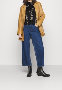 Manteau en cuir marron porté sur un haut noir à motif floral, associé à un jean large en denim bleu et des bottines à talons noires. Silhouette épurée et décontractée.