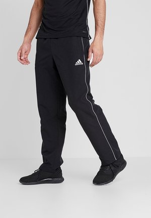 Mann trägt schwarze Adidas Trainingshose mit weißer Paspelierung, schwarze Sneakers und schwarzes T-Shirt, steht vor schlichtem hellgrauem Hintergrund.