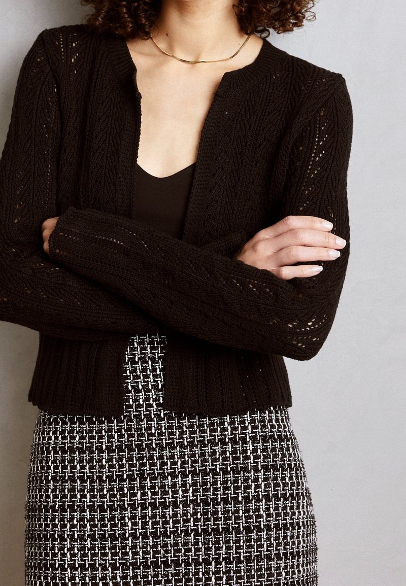 Czarny dziany kardigan z teksturowanym wzorem, ściągaczami na mankietach i krótkim fasonem. Połączony z czarnym topem i kratkowaną spódnicą.