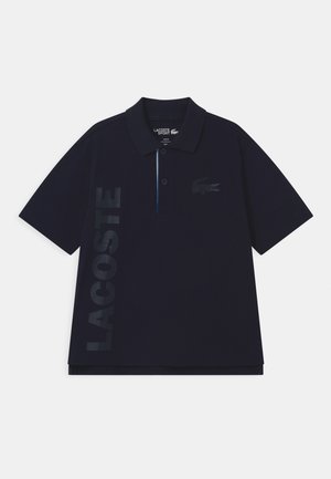 Polo Lacoste Sport bleu marine à manches courtes, patte de boutonnage à deux boutons, grand texte vertical "LACOSTE" et logo crocodile sur la poitrine.