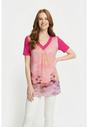 Lächelnde rothaarige Frau, die eine pinke V-Ausschnitt-Tunika mit Palmen- und Wassermotiv trägt, kombiniert mit weißen Hosen, vor weißem Hintergrund stehend.