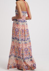 Femme portant une robe maxi sans bretelles et sans dos, avec un motif floral et géométrique multicolore, nouée avec une sangle halter rose.