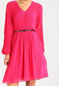 Růžové plisované šaty s dlouhými rukávy, výstřihem do V, zúženým pasem s černým opaskem se cvočky, splývavá sukně, decentní lesk a délka po kolena.