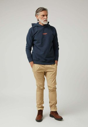 Hombre con cabello y barba canosos, vistiendo una sudadera con capucha azul marino, pantalones beige y botas marrones, de pie con las manos en los bolsillos frente a un fondo liso.