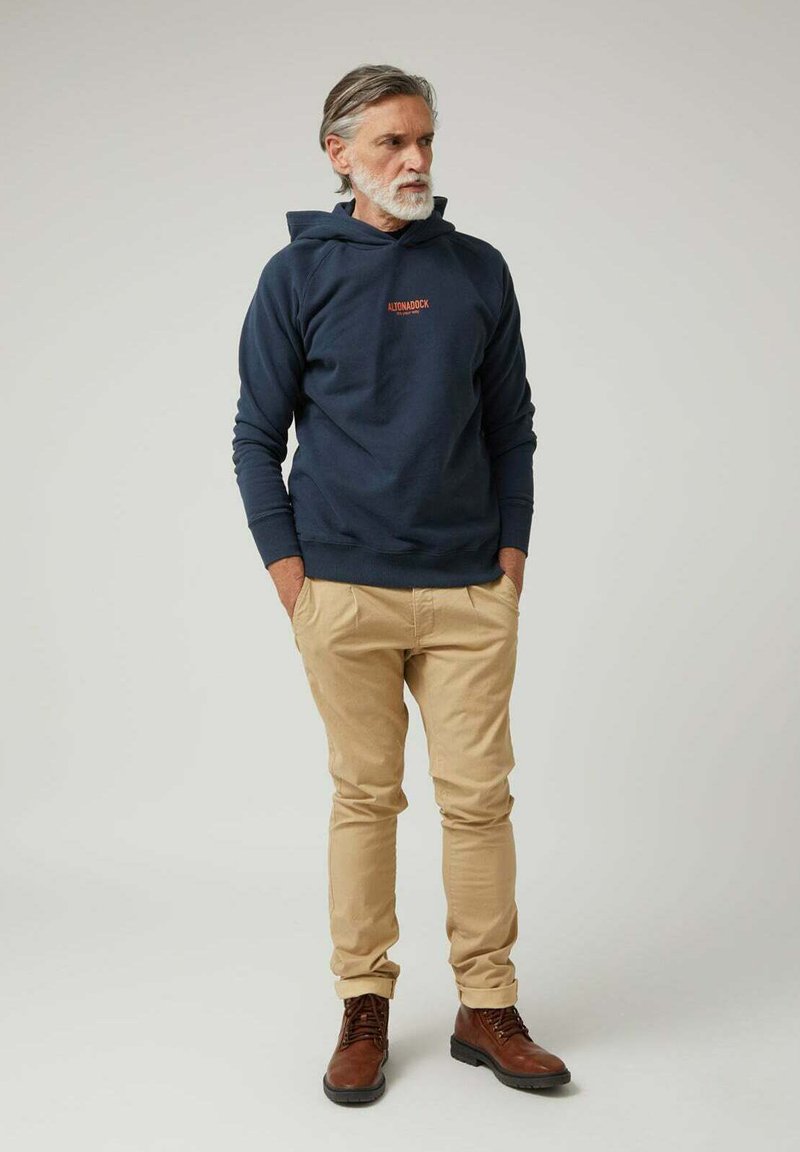 Hombre con cabello y barba canosos, vistiendo una sudadera con capucha azul marino, pantalones beige y botas marrones, de pie con las manos en los bolsillos frente a un fondo liso.