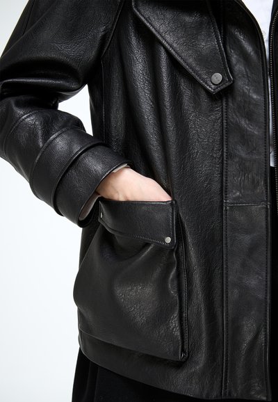 Mano en el bolsillo de una chaqueta de cuero negra que presenta un gran bolsillo con solapa, botones a presión y cierre de cremallera en el lateral.