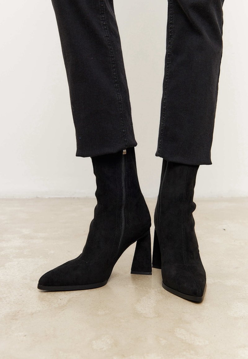 Black suede ankle boots with a pointed toe and chunky heel. Side zipper detail and soft texture; paired with black cropped trousers.