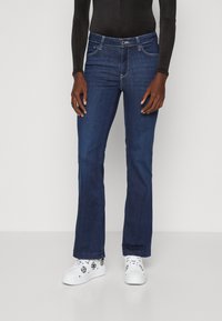 Tummansiniset levenevät farkut etupainikkeella, joissa on viisi taskua ja kontrastiväriset ompeleet. Yhdistetty valkoisiin tennareihin.