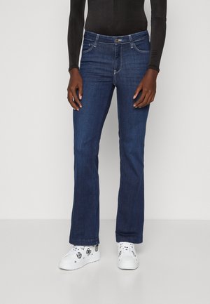 Donkerblauwe, uitlopende denim jeans met een sluiting aan de voorkant met knoop, vijf zakken en contrasterende stiknaad. Gekozen in combinatie met witte sneakers.