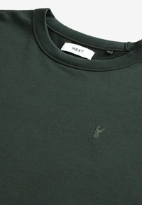 T-shirt vert foncé col rond avec un petit logo tête de cerf brodé sur la poitrine et une étiquette blanche de la marque NEXT à l'intérieur du col.