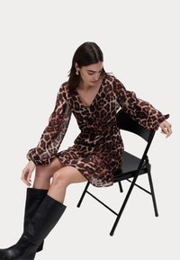 Robe à imprimé léopard avec des manches longues en voile, décolleté en V et jupe évasée, associée à des bottes noires jusqu'aux genoux, assise sur une chaise pliante.
