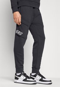Mörkgrå joggers med cargofickor, med en vit logotyp på låret. Bärs med svartvita sneakers, vilket förstärker en sportig look.