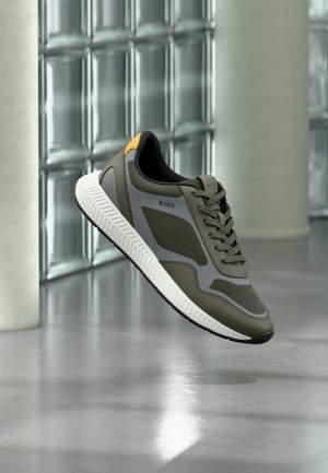 Buty sportowe BOSS w oliwkowo-zielonym i szarym kolorze z białą podeszwą i żółtym detalem na pięcie, unoszące się nad gładką betonową podłogą przy ścianie z bloczków szklanych.