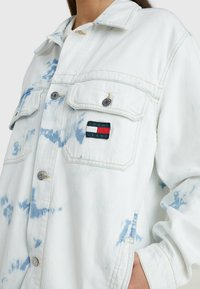 Weiße Denimjacke mit blauen Batikmustern, zwei Brusttaschen, Knopfverschluss und aufgesticktem "TOMMY JEANS" Logo-Patch.