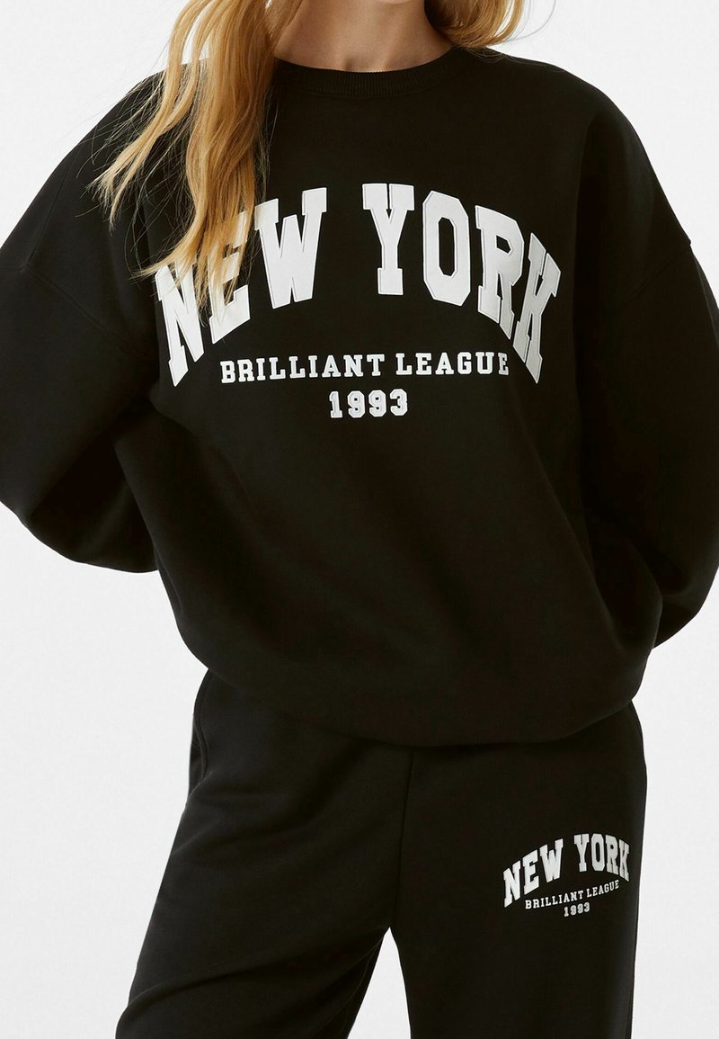 Ensemble sweat-shirt et pantalon noirs avec le texte blanc "NEW YORK Brilliant League 1993" porté par une personne aux longs cheveux blonds.