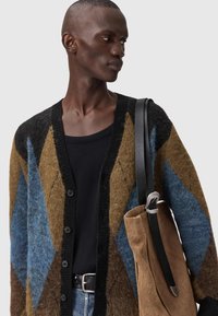 Πολύχρωμη πλεκτή ζακέτα με γεωμετρικό σχέδιο σε καφέ, μπλε και μαύρο, με εμπρόσθια κουμπιά. Το μοντέλο φοράει μαύρο τοπ και κρατά μια τσάντα.