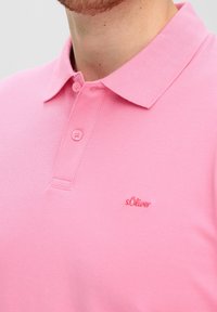 Růžová polo košile s měkkou texturou, klasickým límcem a dvěma předními knoflíky. Název značky "s.Oliver" je vyšitý červenou barvou.